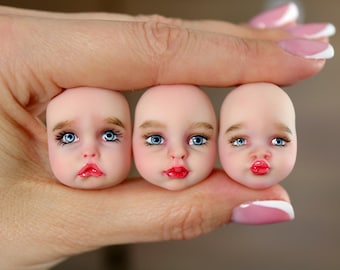 May include: Three miniature doll heads with pale skin tones, light brown eyebrows, and bright blue eyes. Each head features distinct facial expressions, including a sad mouth, a neutral mouth, and a puckered mouth, all with painted red lips.