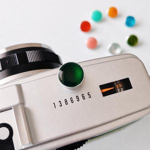 Peut inclure: Un capot supérieur d'appareil photo argenté avec un déclencheur circulaire vert foncé, le numéro de série 1386965 et une petite fenêtre affichant une aiguille orange pointant entre 20 et 22. Sept petits cabochons colorés sont à proximité.