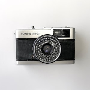 May include: An Olympus Trip 35 film camera with a silver metal body and black textured grip. The front features a circular lens with a 40mm focal length and a 1:2.8 aperture, surrounded by a silver ring with text detailing the lens specifications.