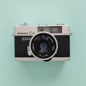 May include: A silver and black Konica C35 film camera featuring a Hexanon 38mm 1:2.8 lens. The front body displays the text Konica C35 and flashmatic. The lens barrel includes markings for DIN, ASA, and focal length, with a textured black grip.