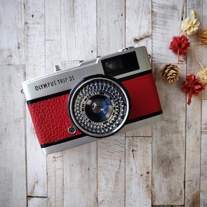 May include: A vintage Olympus Trip 35 camera with a red leatherette body and silver accents. The camera lens is black with silver detailing. The text "OLYMPUS TRIP 35" is printed on the top. The camera is on a light wood surface.
