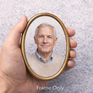 Op de afbeelding: Een ovale, goudkleurige fotolijst met een portret van een glimlachende oudere man. Het frame wordt in een hand gehouden tegen een lichtgrijze achtergrond. De tekst "Frame Only" staat onderaan de afbeelding.