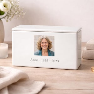 May include: A white rectangular cremation urn with a photograph of a woman and the inscription "Anna 1956-2023." The urn is on a wooden surface, with a lit candle, flowers, and a necklace in the background. A memorial item.
