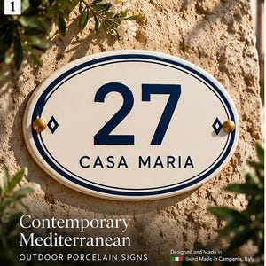 Peut inclure: Une plaque de maison ovale en porcelaine avec un fond crème et une bordure bleu foncé. La plaque affiche le numéro 27 en grands chiffres bleu foncé au-dessus du texte CASA MARIA en lettres bleu foncé, fixée par deux boulons dorés.