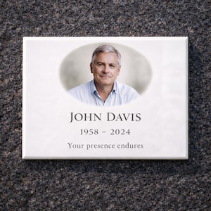Könnte beinhalten: Eine weiße Gedenktafel mit einem Porträt eines Mannes, dem Namen "JOHN DAVIS" und den Jahren "1958 - 2024". Der Text "Your presence endures" steht darunter. Die Tafel befindet sich vor einem strukturierten, dunkelgrauen Hintergrund.