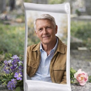Puede incluir: Marco conmemorativo de cerámica blanca, en forma de pergamino, con una foto de un hombre mayor sonriendo, chaqueta marrón y camisa azul. Flores moradas y una rosa rosa sobre una superficie de piedra.