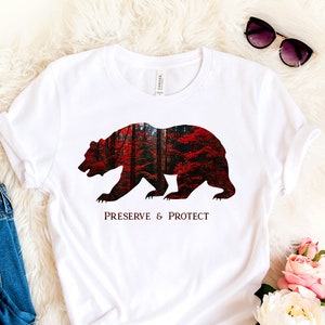 May include: White t-shirt featuring a black bear silhouette. The bear is filled with a red and orange forest scene. The text "Preserve & Protect" is printed below the bear.