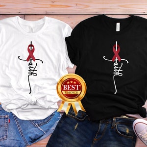 Puede incluir: Dos camisetas con una cruz blanca y la palabra "fe" escrita verticalmente. Una cinta roja está atada alrededor de la cruz. Una camiseta es blanca y la otra es negra.