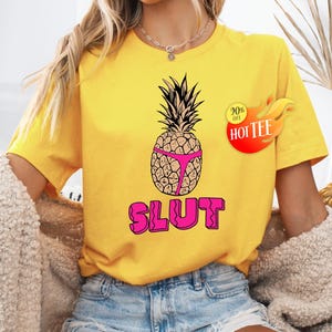 Pode incluir: Camiseta amarela com um design de abacaxi usando roupa íntima rosa. A palavra "SLUT" é impressa em letras rosa abaixo. Um adesivo vermelho e amarelo com o texto "20% off HOT TEE" está no lado direito.