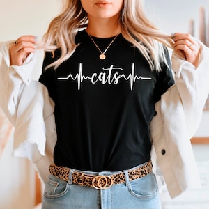 Może przedstawiać: Czarny t-shirt z białym nadrukiem w kształcie bicia serca z napisem "cats" w środku. Osoba nosi białą kurtkę i pasek w panterkę.