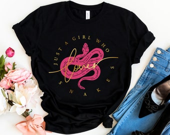 Puede incluir: Camiseta negra con un gráfico de serpiente rosa y texto dorado que dice "Just a girl who loves snakes".