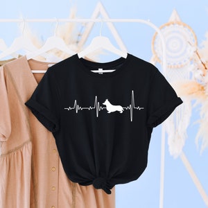 May include: Black t-shirt with a white silhouette of a dog with a heartbeat line above it. The text on the shirt reads "My heart beats for a Corgi".