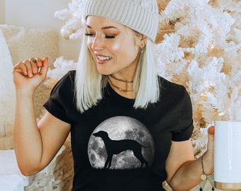 May include: A black t-shirt with a graphic of a dog silhouette on a moon. The dog is facing right and the moon is gray with a white outline. The person wearing the shirt is sitting in front of a white Christmas tree.