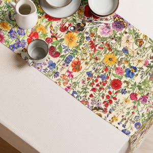 May include: A floral table runner with a cream background and a vibrant pattern of colorful wildflowers, including red poppies, yellow sunflowers, and blue cornflowers. The runner is placed on a white tablecloth, with tableware visible in the background.