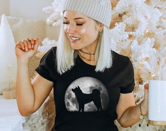 Pode incluir: Uma camiseta preta com a silhueta de um cão em pé em frente a uma lua cheia. O cão é preto e a lua é branca. A camiseta está sendo usada por uma pessoa com cabelo loiro e um gorro branco.