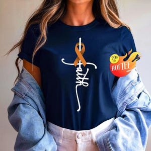 May include: Navy blue t-shirt featuring a white cross with an orange ribbon and the word "faith" in a script font. A promotional sticker with the text "20% off HOT TEE" is visible on the right side. The shirt is a casual wear item.