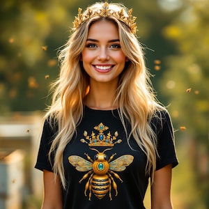以下が含まれることがあります： 金色の蜂と王冠のデザインが施された黒いTシャツを着た女性。蜂は体にターコイズの宝石を付けています。彼女は金色の王冠をかぶって微笑んでいます。