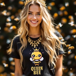 以下が含まれることがあります： 黒いTシャツを着た女性。Tシャツには、漫画の蜂のイラストと「Queen Bee」の文字がプリントされています。彼女は金の王冠と金のネックレスをしています。背景はぼやけていて、蜂の群れが写っています。