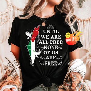 以下が含まれることがあります： 黒いTシャツ。パレスチナの国旗の色で描かれたパレスチナの地図、鳩、そして「UNTIL WE ARE ALL FREE NONE OF US ARE FREE」という文字がプリントされています。「Hot Tee」のステッカーも表示されています。