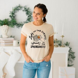 May include: A light beige t-shirt with a black graphic print that reads "Lady in the streets, freak in the spreadsheets".