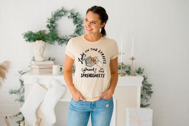 May include: A light beige t-shirt with a black graphic print that reads "Lady in the streets, freak in the spreadsheets".