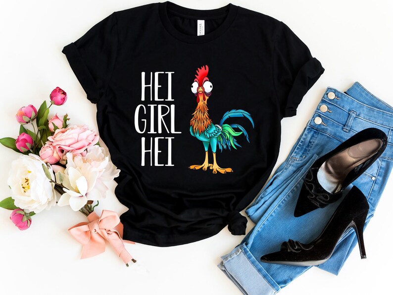 Puede incluir: Camiseta negra con un gr&aacute;fico blanco de un gallo con el texto "Hei Girl Hei". La camiseta est&aacute; colocada sobre una superficie blanca con un ramo de flores, un par de pantalones vaqueros azules y tacones negros.
