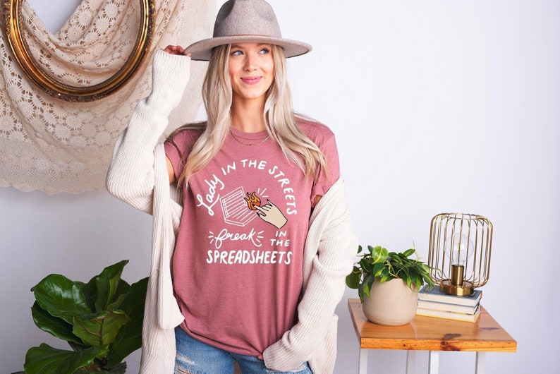 May include: A mauve t-shirt with a white graphic that says "Lady in the streets, freak in the spreadsheets" with a drawing of a hand holding a burning spreadsheet.