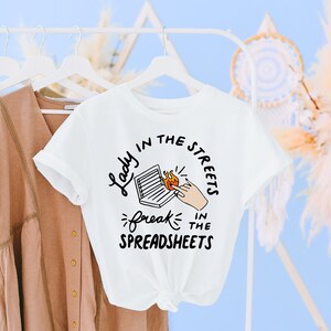 May include: White t-shirt with a graphic design that says "Lady in the streets, freak in the spreadsheets". The graphic features a laptop with a hand reaching towards a flame.