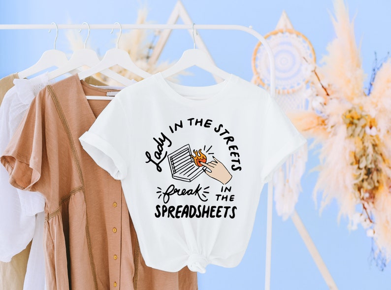 May include: White t-shirt with a graphic design that says "Lady in the streets, freak in the spreadsheets". The graphic features a laptop with a hand reaching towards a flame.