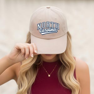 May include: A beige baseball cap featuring the text "NORTH Carolina" in blue and white. The cap is held up by a person with long blonde hair and a red top.