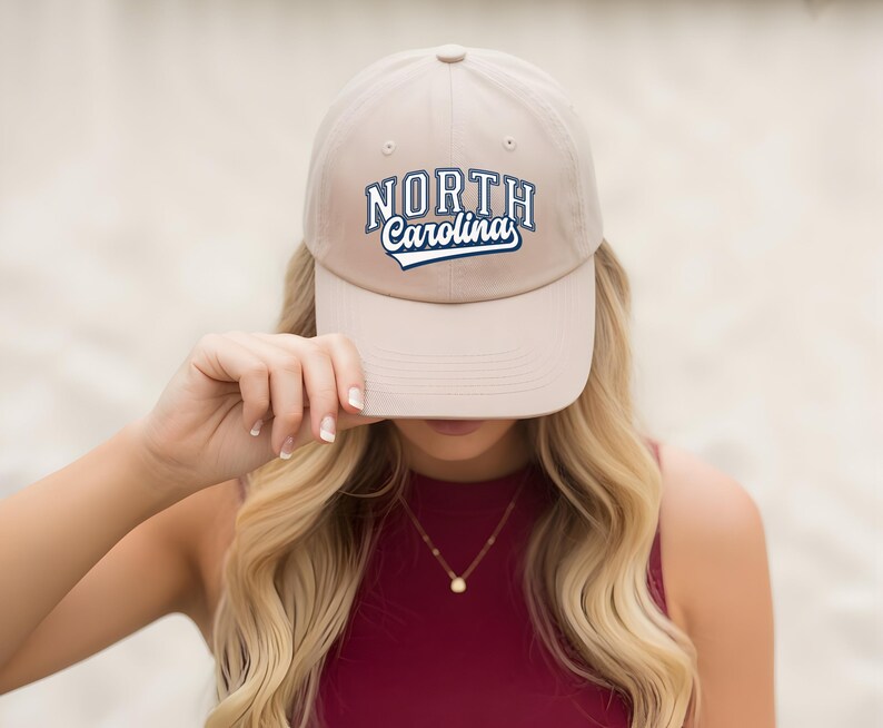 May include: A beige baseball cap featuring the text "NORTH Carolina" in blue and white. The cap is held up by a person with long blonde hair and a red top.