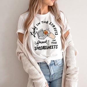 May include: White t-shirt with black text that reads "Lady in the streets, freak in the spreadsheets". The text is surrounded by a circle and includes a drawing of a hand holding a laptop on fire.