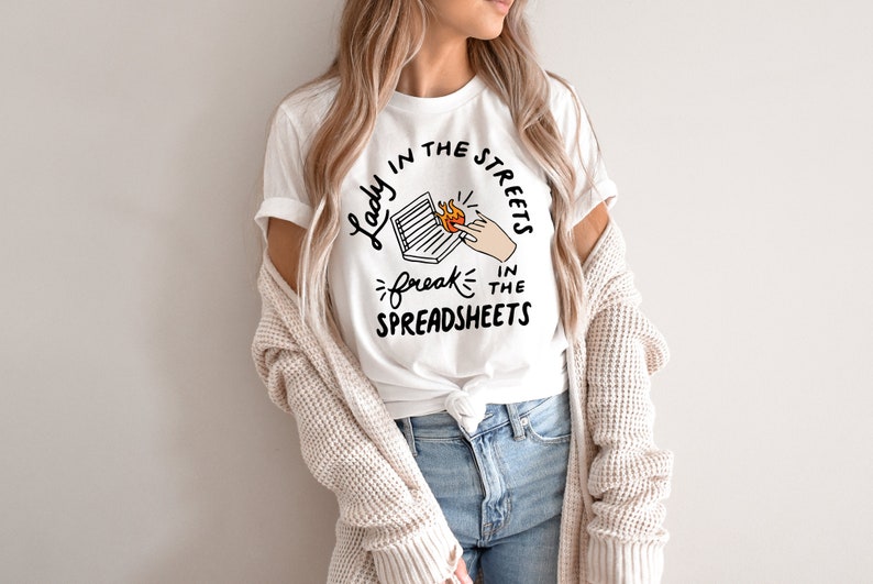 May include: White t-shirt with black text that reads "Lady in the streets, freak in the spreadsheets". The text is surrounded by a circle and includes a drawing of a hand holding a laptop on fire.