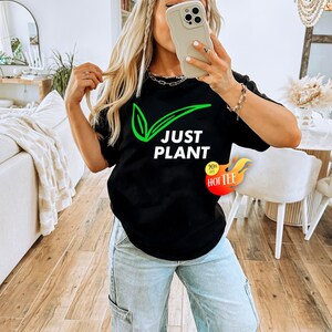 May include: Black t-shirt with the text "JUST PLANT" in white and a green leaf design. The shirt also has a "90% off HOT TEE" sticker. The person in the image is wearing light blue jeans.