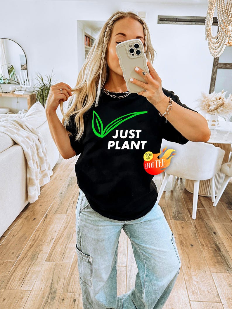 May include: Black t-shirt with the text "JUST PLANT" in white and a green leaf design. The shirt also has a "90% off HOT TEE" sticker. The person in the image is wearing light blue jeans.