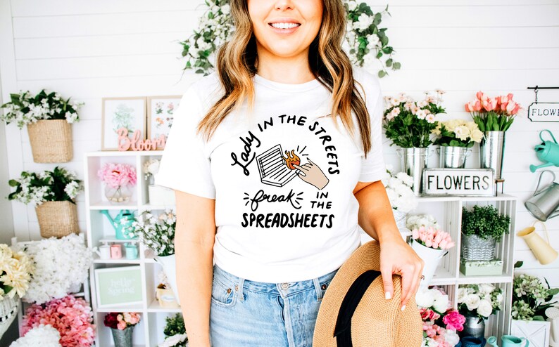 May include: White t-shirt with a black graphic design that says "Lady in the Streets, Freak in the Spreadsheets". The graphic features a laptop with flames coming out of it and a hand pointing at the flames.