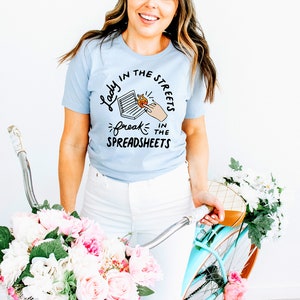 May include: A light blue t-shirt with a graphic design that says "Lady in the streets, freak in the spreadsheets". The graphic features a hand reaching into a spreadsheet with a flame coming out of it.