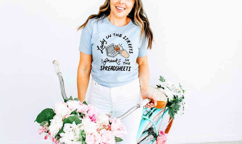 May include: A light blue t-shirt with a graphic design that says "Lady in the streets, freak in the spreadsheets". The graphic features a hand reaching into a spreadsheet with a flame coming out of it.