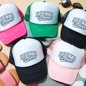 May include: A selection of trucker caps in shades of pink, green, black, and white. Each cap displays the text "NORTH Carolina" in a decorative font. The caps have a mesh back and a curved peak.