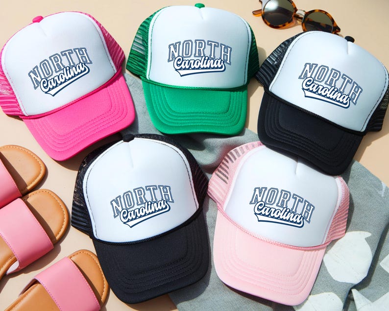 May include: A selection of trucker caps in shades of pink, green, black, and white. Each cap displays the text "NORTH Carolina" in a decorative font. The caps have a mesh back and a curved peak.