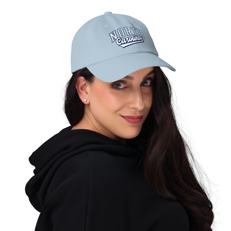 May include: Pale blue baseball cap featuring the text "North Carolina" in white and navy blue. The cap has a curved peak and an adjustable strap. The hat is made of a soft, durable material.