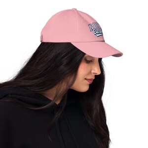 May include: A pale pink baseball cap with a curved peak. The cap displays the text "North Carolina" in a blue and white design on the front. The cap is made of a soft material and has an adjustable strap.