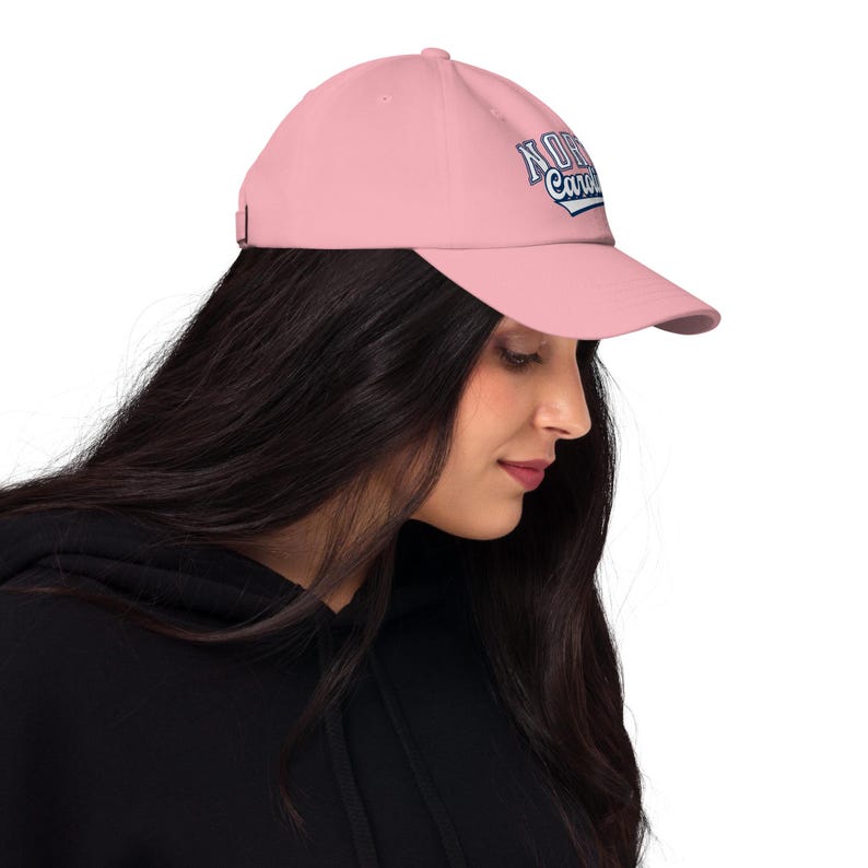 May include: A pale pink baseball cap with a curved peak. The cap displays the text "North Carolina" in a blue and white design on the front. The cap is made of a soft material and has an adjustable strap.