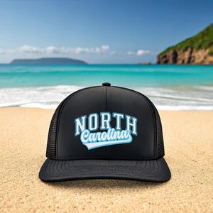 May include: Black trucker hat with the text "NORTH Carolina" in white and light blue. The hat is on a sandy beach with the ocean and sky in the background.