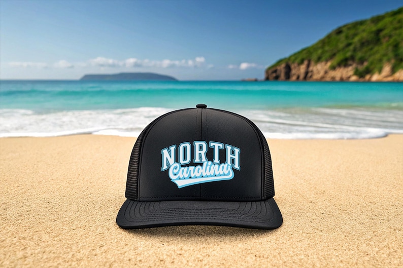 May include: Black trucker hat with the text "NORTH Carolina" in white and light blue. The hat is on a sandy beach with the ocean and sky in the background.