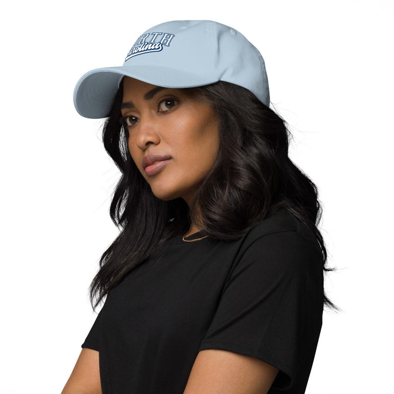 May include: A pale blue baseball cap featuring the text "North Carolina" in white and navy blue script. The cap has a curved peak and a classic six-panel design. The hat is made of a soft, durable material.