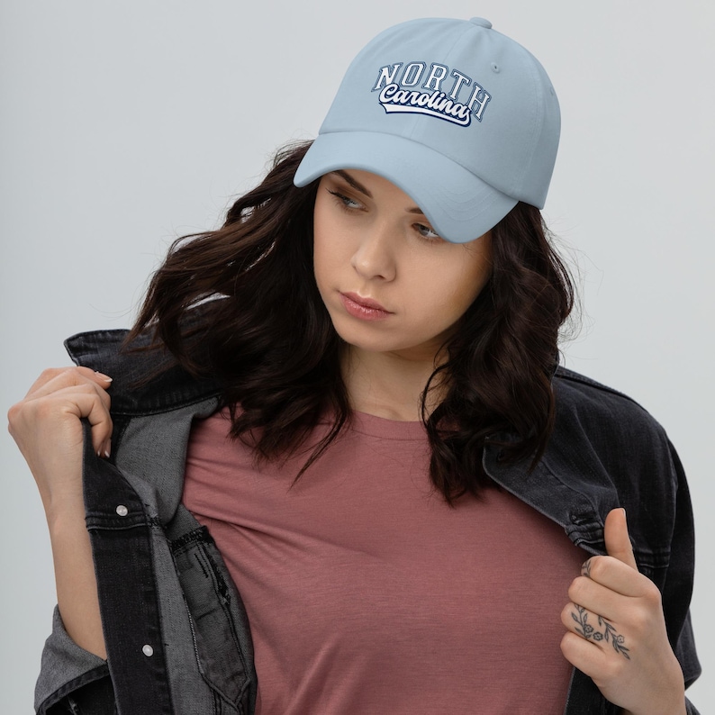 May include: Pale blue baseball cap featuring the text "NORTH Carolina" in a white and navy blue design. The cap is made of a soft material and has a curved peak. A classic style hat.