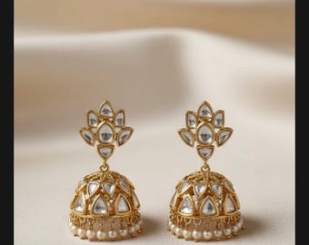 May include: A pair of gold-colored, ornate earrings. Each earring features a floral design at the top and a bell-shaped bottom. The bell-shaped portion is adorned with clear, triangular-shaped stones and small, white pearls along the bottom edge. The earrings are set against a neutral background.