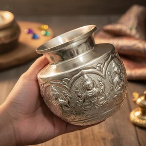 May include: A silver-colored metal pot with intricate embossed designs featuring figures. The pot has a wide, flared rim and a rounded body. The detailed carvings and polished finish give it a decorative appearance.