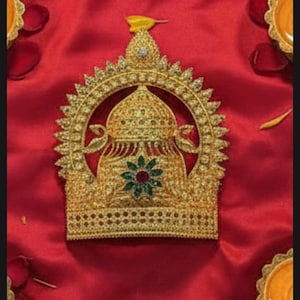 May include: A gold-colored crown with intricate detailing, set against a red satin background. The crown features a central floral design with green and red accents, surrounded by a decorative arch and a pointed top.